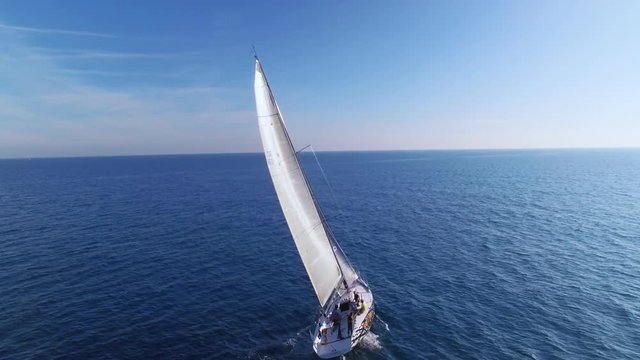 Aerial drone follows beautiful luxurious competition and racing yacht or sailboat on journey or cruise through mediterranean sea or ocean, with spinnaker and mainsail full of wind