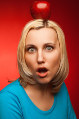 Portrait of shocked beautiful student girl posing over red background with red apple on head. Education, Girl next door concept. Perfect blond hair, natural make-up. Hipster style. Close up