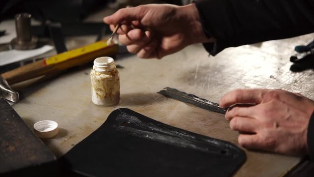 Leathersmith is applying glue on a leather parts of the zipper with a special brush. He works at the table in the workshop.