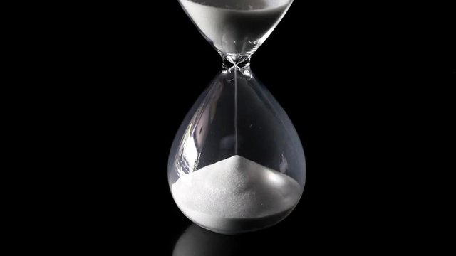 Falling sand showing the passing time of hourglass close-up and isolated on glossy black floor and background. Presented in the centre of the screen.