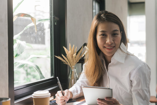 Young business woman in white dress sitting at table in cafe and writing in notebook. Asian woman using tablet and cup of coffee. Freelancer working in coffee shop. Student learning online.