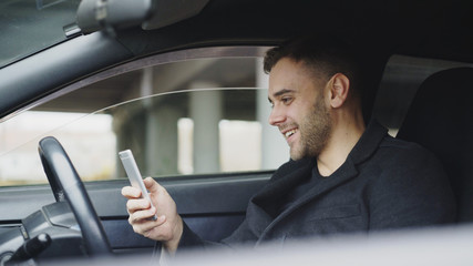 Attractive businessman sitting inside car laughing while using smartphone after trip