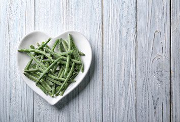 Heart-shaped plate with frozen green beans on wooden background