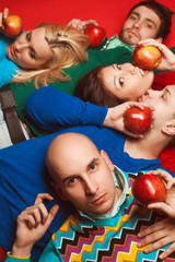 Fototapeta na wymiar Picnic concept. Portrait of four stylish close friends hugging, having rest, holding red apples and lying on red background. Guys having fun. Hipster style. Studio shot