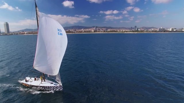 Aerial footage of drone flying around beautiful racing competition yacht or sailboat, with team of sailors on deck preparing entrance into marina, spinnaker up with amazing sun flare