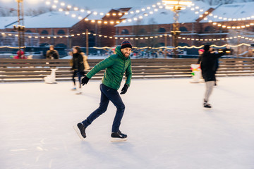 Obraz premium Joyful bearded man practices going skating on ice ring, has cheerful expression, smiles happily, demontrates his professionalism. Active sporty male in green jacket go in for winter sport
