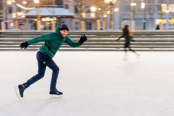 Happy active sporty male involved in winter activities, demonstrates his skating skills on Christmas decorated ice ring, being in movement, has active lifestyle, enjoys his favourite hobby