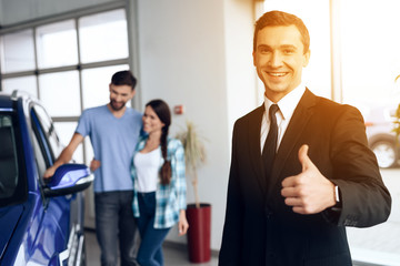 A car salesman is posing for a camera near buyers who are choosing a new car.