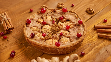 Tasty apple pie with fresh red cranberries and walnuts decorated with ginger and sticks of cinnamon. Just backed apple pie ready to eat on wooden brown background. Side view