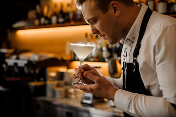 Young barman with closed eyes holding a glass of fresh alcoholic cocktail