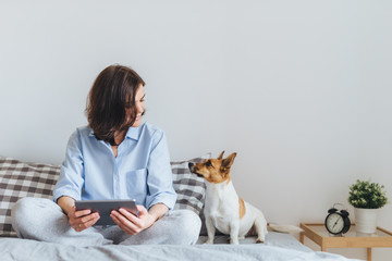 Beautiful brunette female in pyjamas sits on bed in bedroom with her jack russell terrier dog,...
