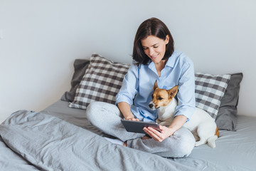 Domestic cozy atmosphere. Beatiful female model wears pyjmas, sits on bedclothes in bedroom with her favourite pet, messages with frineds via tablet computer, enjoys free internet connection