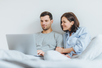 Affectionate female and male have joy together, watch film in bedroom on laptop computer, enjoy togetherness and free internet connection. Lovely couple in love have good time in bedroom
