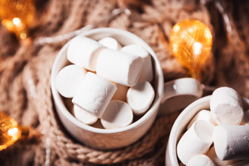 Christmas background with cocoa or coffee with marshmallows in a white cup on a brown knitted winter scarf and a glowing golden garland. Beautiful concept of home comfort and warmth