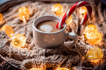 Christmas background coffee in a white cup with candy on a brown knitted winter scarf and a glowing golden garland. Beautiful home Holiday drink