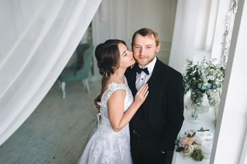 The bride is stretching to kiss her groom on the cheek. Wedding photography in the interior.