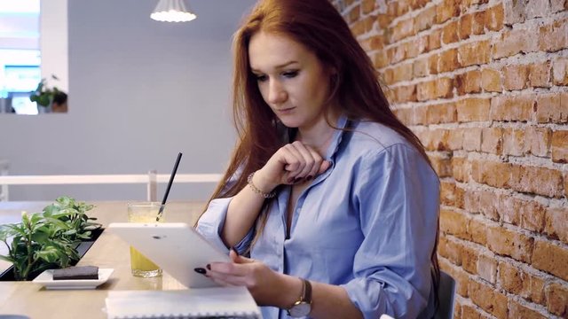 Young, pretty woman with red hair using tablet while sitting in cafe 
