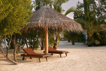 Two beach deck chairs under straw umbrella. Indian ocean coastline on Maldives island. White sandy beach and calm sea. Travel and vacation concept.