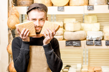 Funny portrait of a handsome cheese seller in uniform standing with seasoned cheese in front of the store showcase full of different cheeses