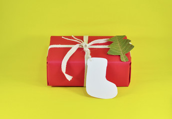 Christmas gift wrapped by red paper tied the box by Light Brown Cotton Twill Tape with white paper tag card sock shape and dry leaf pine tree shape on yellow background
