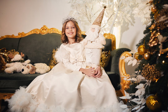 a girl dressed in the image of a snow queen posing in an atmospheric New Year's room with a beautiful Christmas tree, toys and dressed in a white long dress and a silver crown