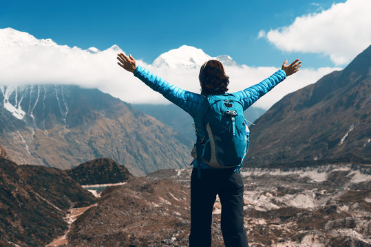 Standing young woman with backpack and raised up arms on the hill and looking on mountains. Landscape with happy girl, mountains, blue sky with clouds in autumn in Nepal. Travel. Trekking in Himalayas
