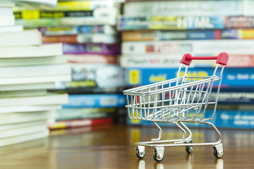 Mini shopping cart on wooden table with blurred pile of book as background, concept of shopping online knowledge to advance