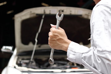 Professional young mechanic man in uniform holding wrench with open hood at the repair garage. Car insurance concept.