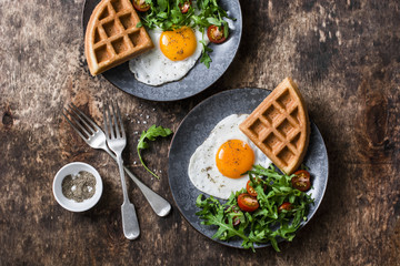 Savory waffles, fried eggs and arugula, cherry tomato salad - delicious healthy breakfast on wooden background, top view