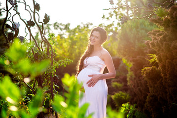 A happy pregnant girl in a white dress is walking in the park.