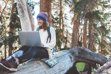 Traveling beautiful woman with backpack and knitted hat using laptop, typing text and sitting on tree in wild forest at sunny day. Smiling.