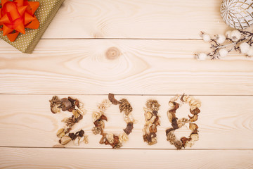 2018 inscription of dried flowers, a golden gift box with an orande bow and white vintage Christmas-tree decorations on a light wooden background. Top view. Copyspace.