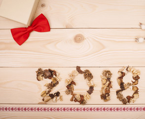 2018 inscription of dried flowers with a beige gift box and a red fabric bow at the top and patterned ribbon at the bottom on a light wooden background. Top view. Copyspace.