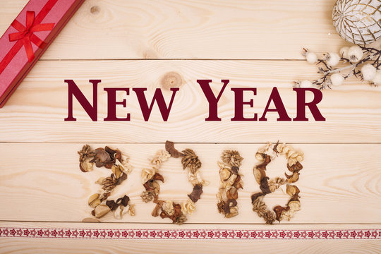 New Year 2018 inscription of dried flowers with a red gift box and white vintage ornaments at the top and patterned ribbon at the bottom on a light wooden background. Top view. Copyspace.