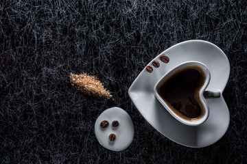 A heart shaped coffee cup with coffee beans and spilled milk and brown sugar on a black and silver kitchen table top in hard, low key light - 183452438