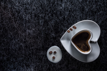 A heart shaped coffee cup with coffee beans and spilled milk on a black and silver kitchen table top in hard, low key light