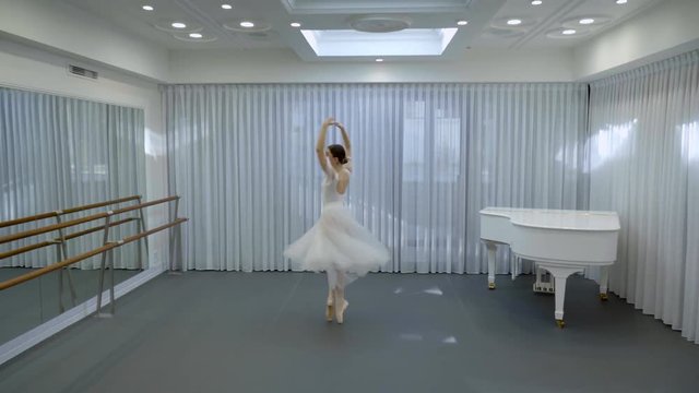Ballerina in long ballet tutu dances in studio with white grand piano. Woman in dance suit and punts whirl in a spacious hall with musical instrument.