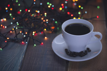 Festive still life with a cup of hot espresso. Coffee beans on a saucer. Fiary Christmas lights in the back. Dark blurred bokeh. Dark wooden background. Close up.