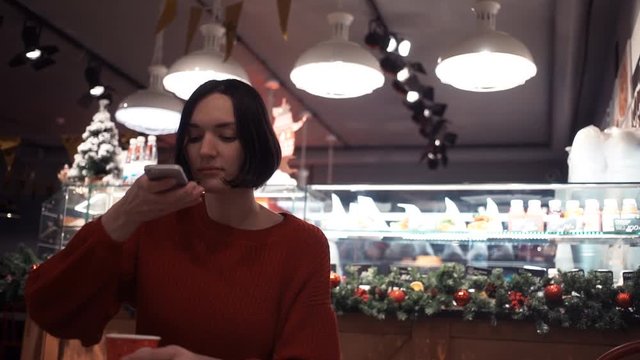 Attractive young womanusing smartphone sitting in cafe. Xmas time. Slow motion