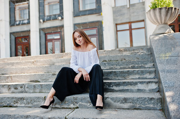 Good-looking young woman in white blouse, wide black pants and black classic high heels sitting on...