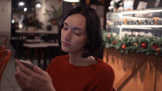 Attractive young girl chatting online using smartphone app sitting in empty cafe