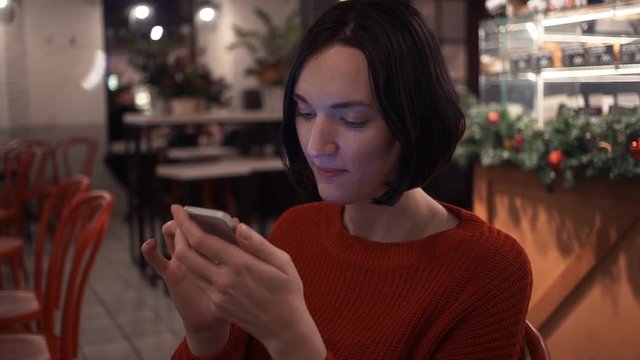 Pretty young girl chatting online using smartphone sitting in cafe in evening