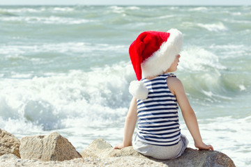 Boy in santa hat sitting on a rock. Sea shore. Back view