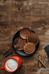 top view of black plate with pancakes and mug of cappuccino on wooden table