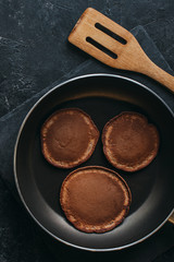 top view of chocolate pancakes on frying pan and wooden spatula on black table