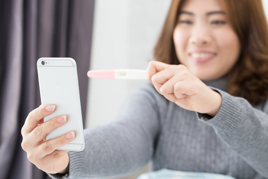 Happy young woman taking selfie by smartphone and holding pregnancy test