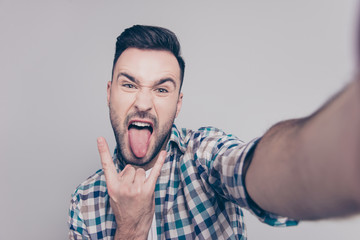 Self portrait of  attractive, crazy man with bristle, stubble, shooting selfiye and showing rock and roll symbol with tongue out to the front camera over grey background
