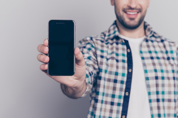 Close up portrait with copy space of black screen of smart phone demonstrating by cheerful, confident, professional, brunet guy with half face in checkered shirt over grey background