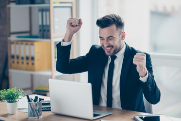 Portrait of young, smiling, attractive, confident finansist in tuxedo, blue jacket, triumphant  having his arms with fists raised, celebrating completed work, sitting at his desk top in work station