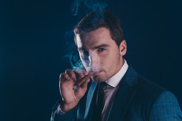 Close up portrait of attractive, elegant, stylish, stunning man smoking cigar over dark blue background